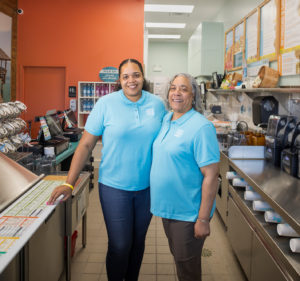 Two women in matching blue shirts stand together in a restaurant kitchen