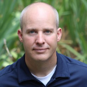 Lance Olson looks into a camera, standing in front of greenery