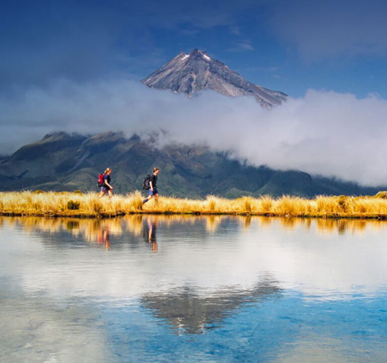 Man and woman wearing backpacks hiking next to a lake with a mountain in the background