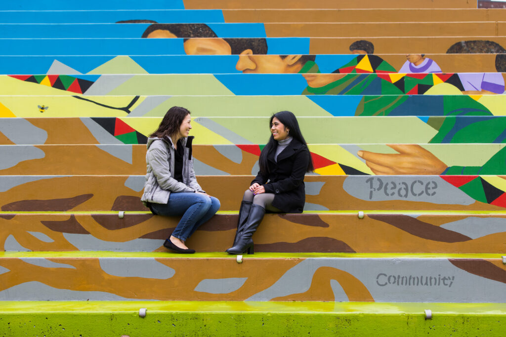 Two women sit outside on stairs while talking