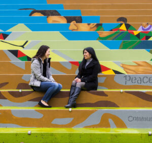 Two women sit outside on stairs while talking