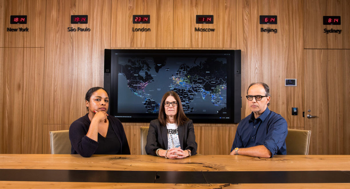 Three people sit at a table with a screen and clocks behind them