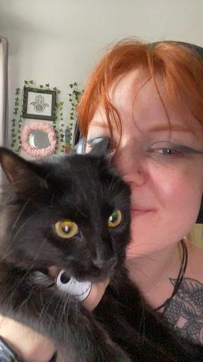 A young woman smiles with her face peering from just behind a cat. 