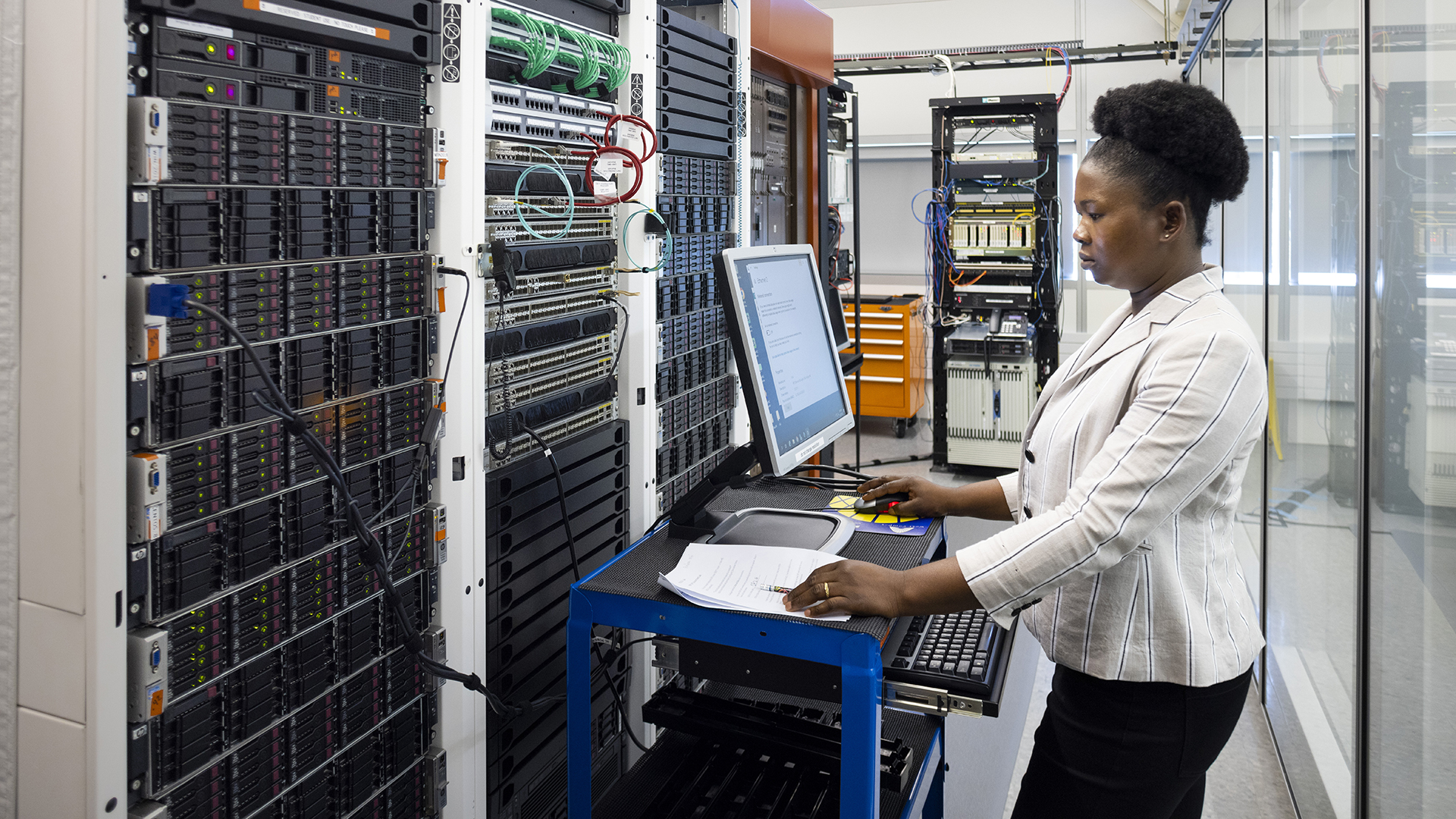 A woman works on a mobile computer station next to a server rack