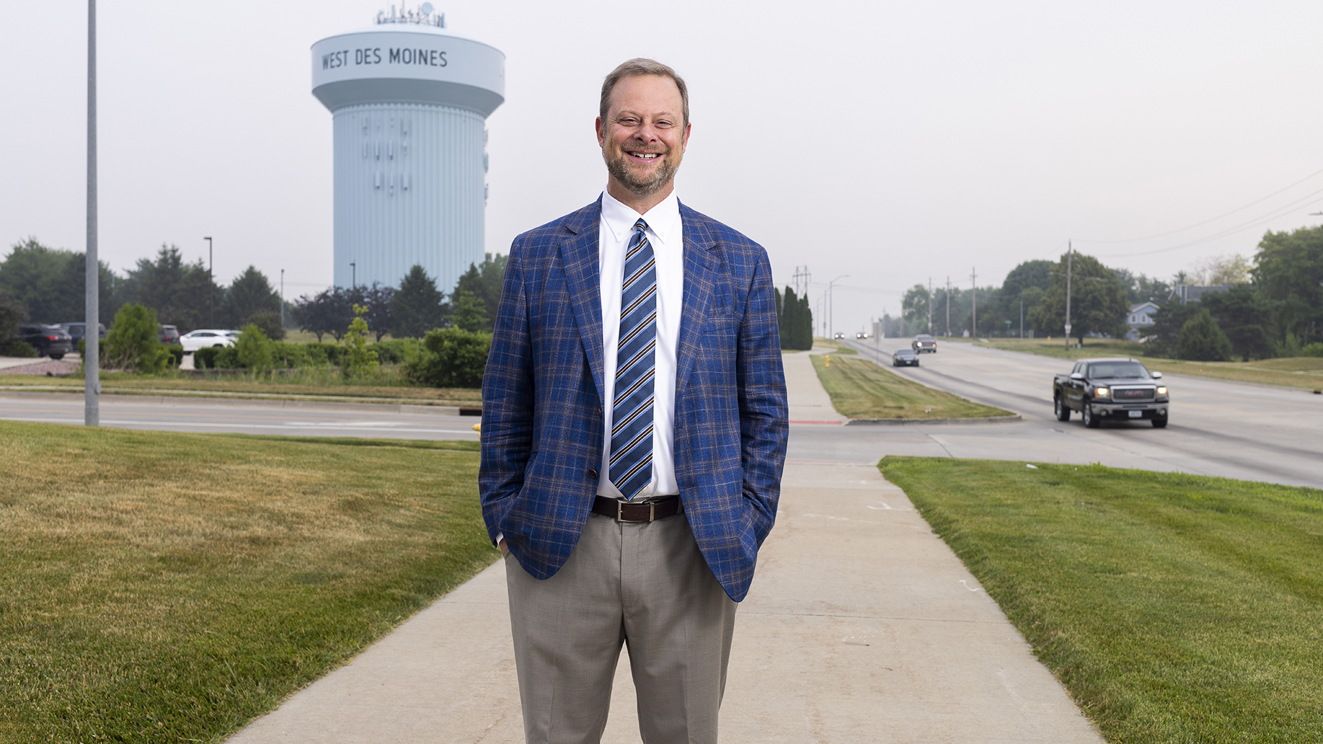 A man in a flannel suit jacket stands on a sidewalk outside with a water tower in the background