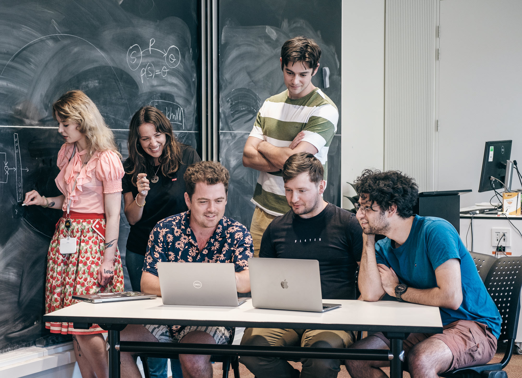 Photo of a group of six young people gathered around a table with computers and a chalkboard in a classroom setting. 