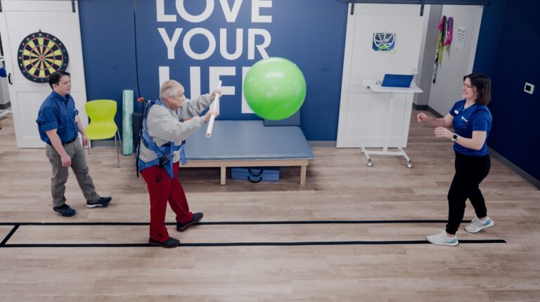 A man wearing a harness attached to the ceiling by a cord catches an exercise ball thrown by a physical therapist.