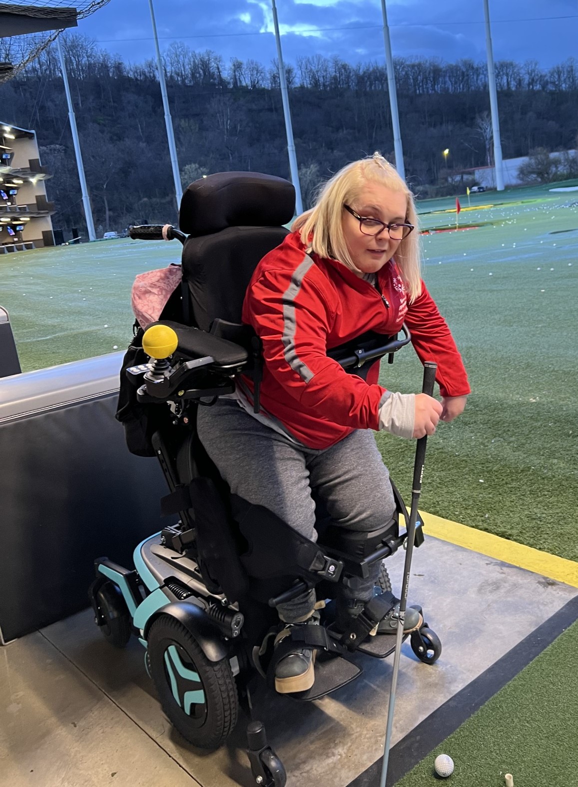 Haleigh Sommers uses a standing brace in her wheel chair to practice hitting gold balls at a driving range.