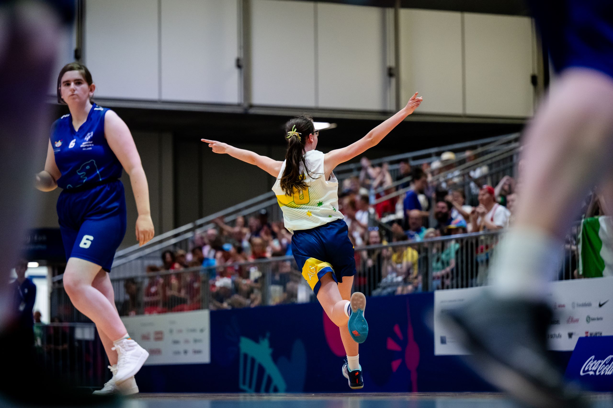 A basketball player, shown from behind, runs with her arms in the air in celebration as a crowd looks on. 