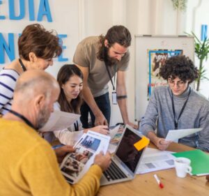 Five people huddle in an office reviewing photos