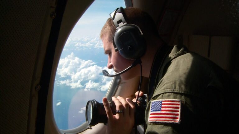 Man in uniform looking out an airplane window, holding a camera