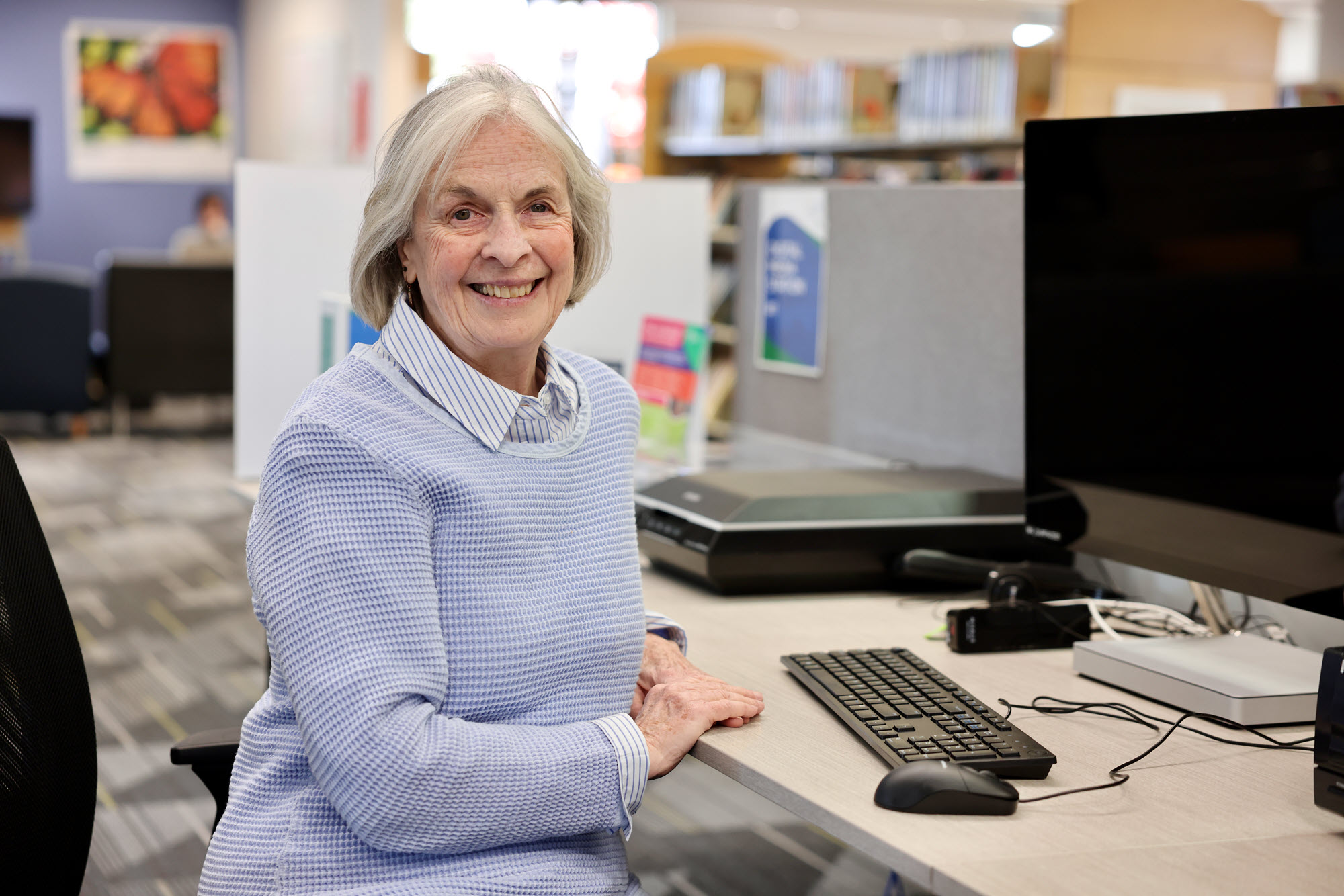 Woman smiling and sitting at a desk with a computer in a library. 