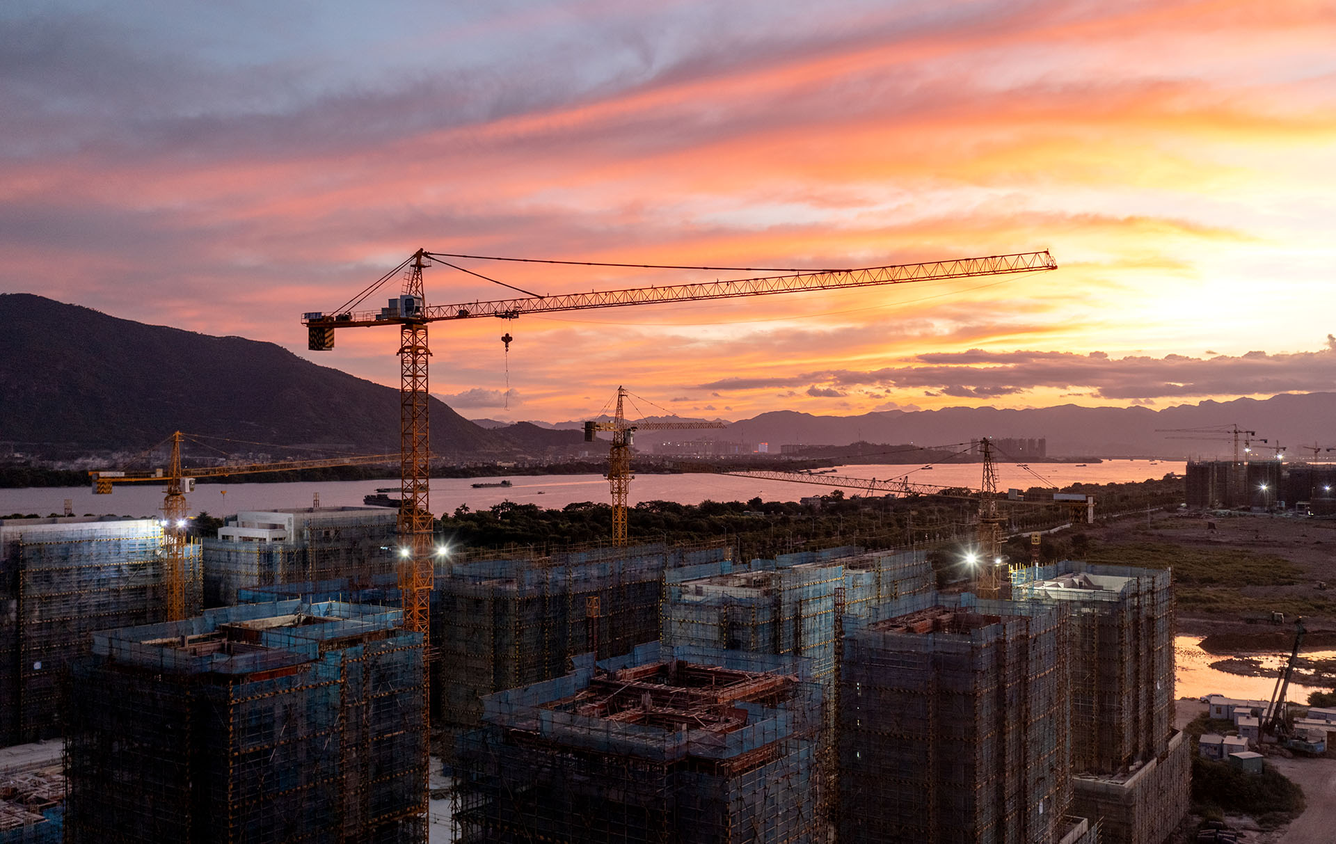 Construction site with cranes towering over partially built structures, illuminated under a sunset sky.