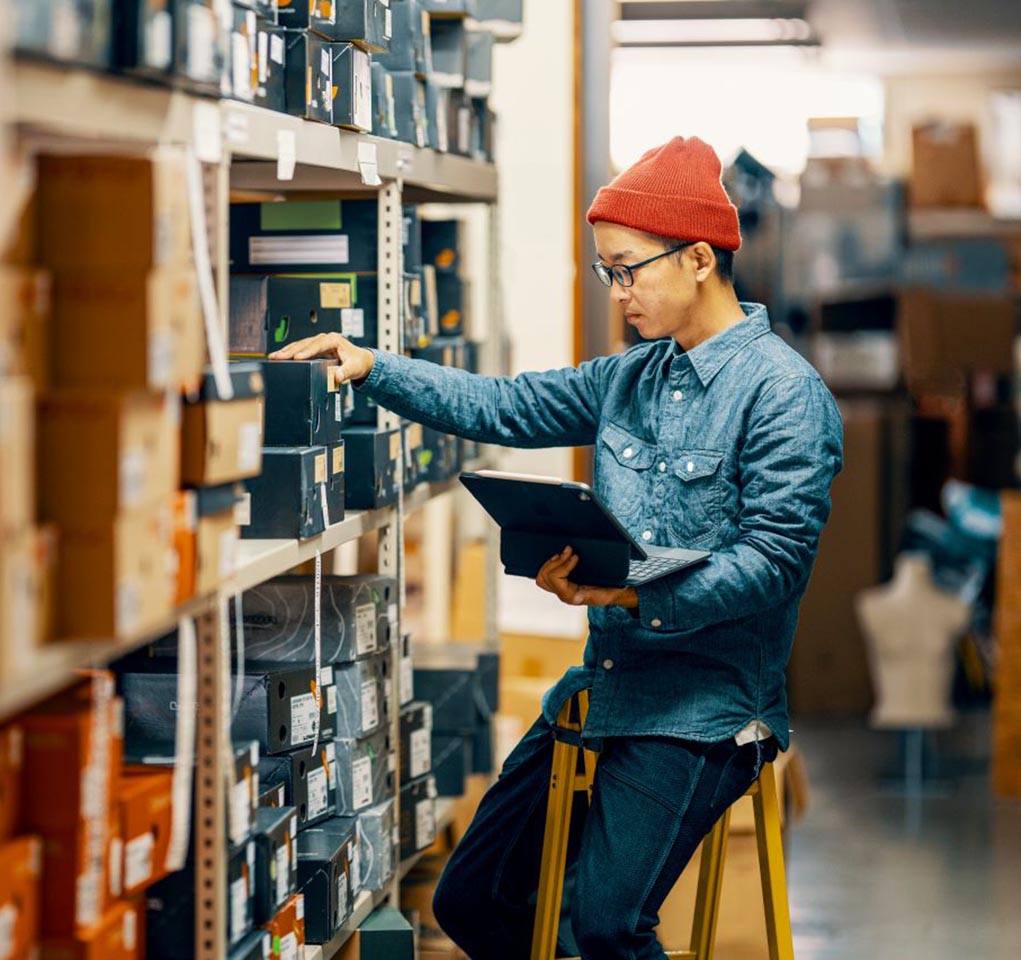 A person sitting on a ladder with a laptop computer inspects inventory in a warehouse