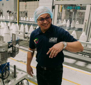 A man in a hairnet and blue polo shirt gestures toward someone out of the frame amid a line of white plastic bottles on a conveyer belt.