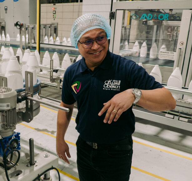 A man in a hairnet and blue polo shirt gestures toward someone out of the frame amid a line of white plastic bottles on a conveyer belt.