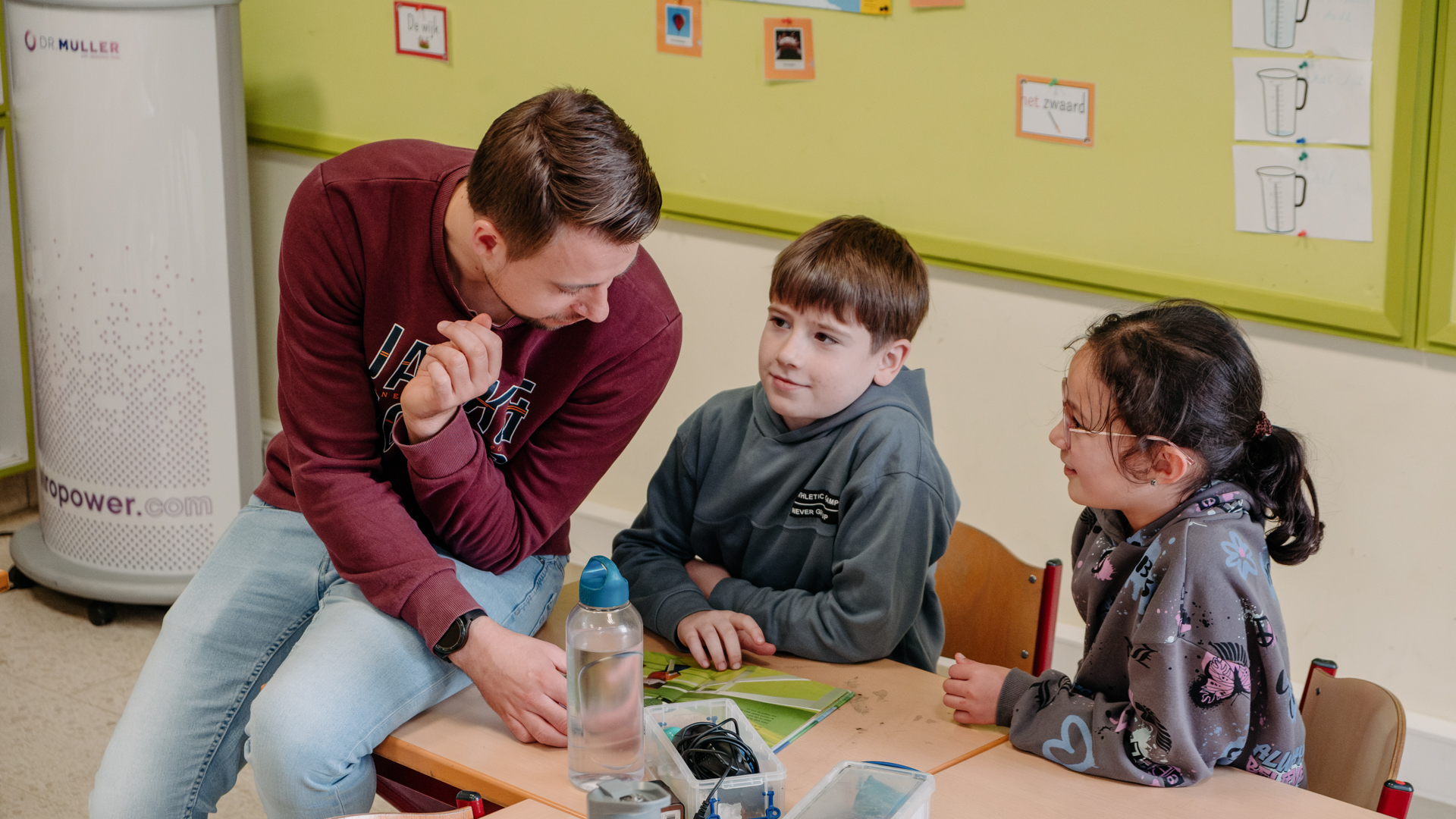 A teacher in a maroon sweatshirt engaging in a discussion with two young students in a classroom setting.