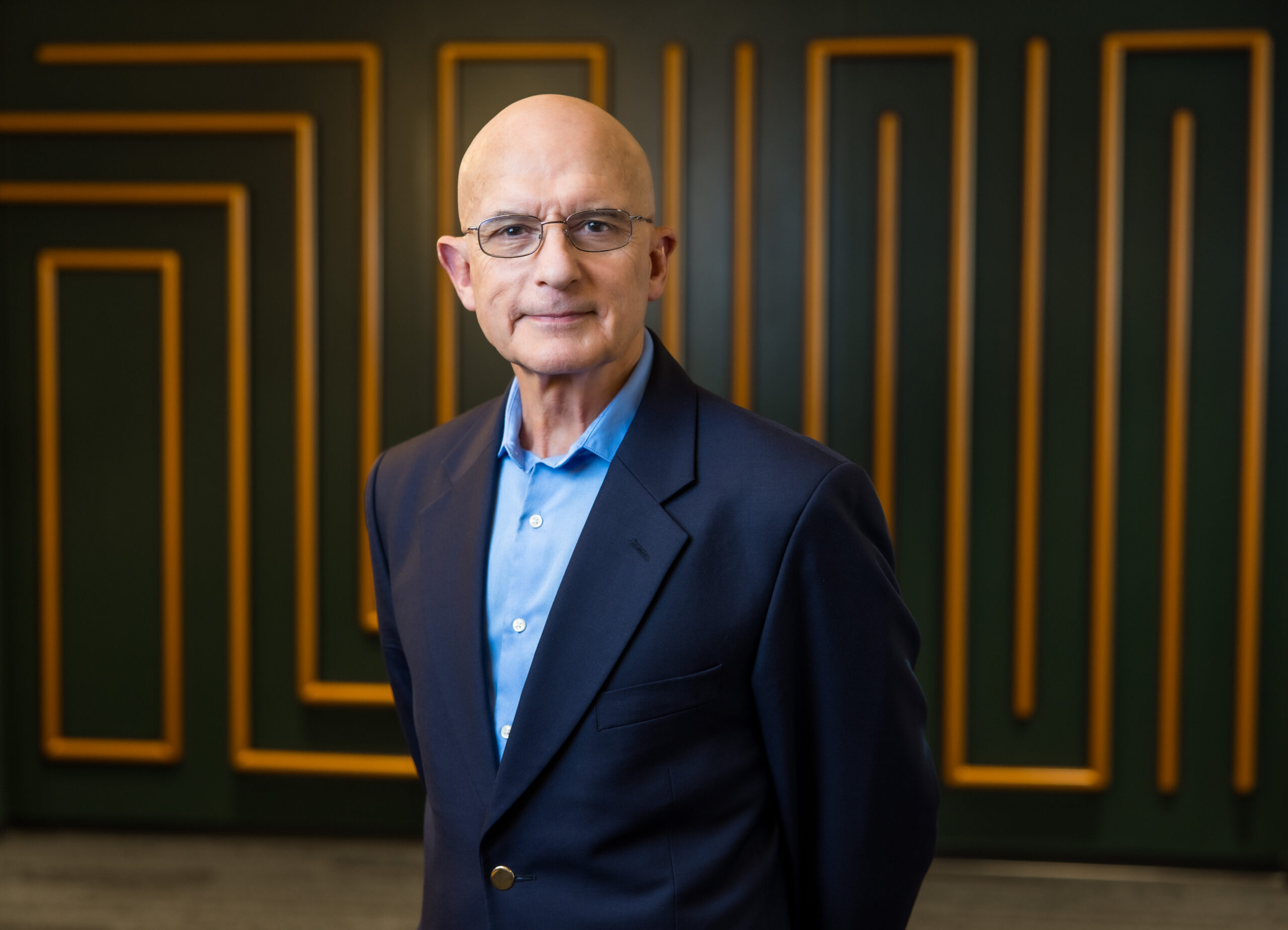 A man wearing glasses, a navy blue blazer, and a light blue shirt, standing in front of a geometric-patterned green and gold wall.