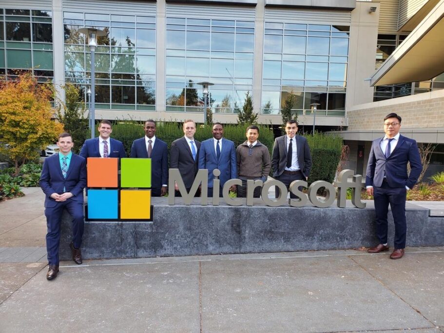 Group of eight people in business attire posing by Microsoft logo sign outside office building.
