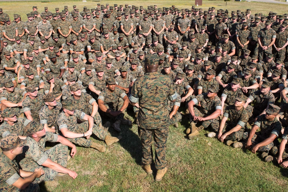 Military personnel in camouflage uniforms seated in formation listening to a standing instructor.